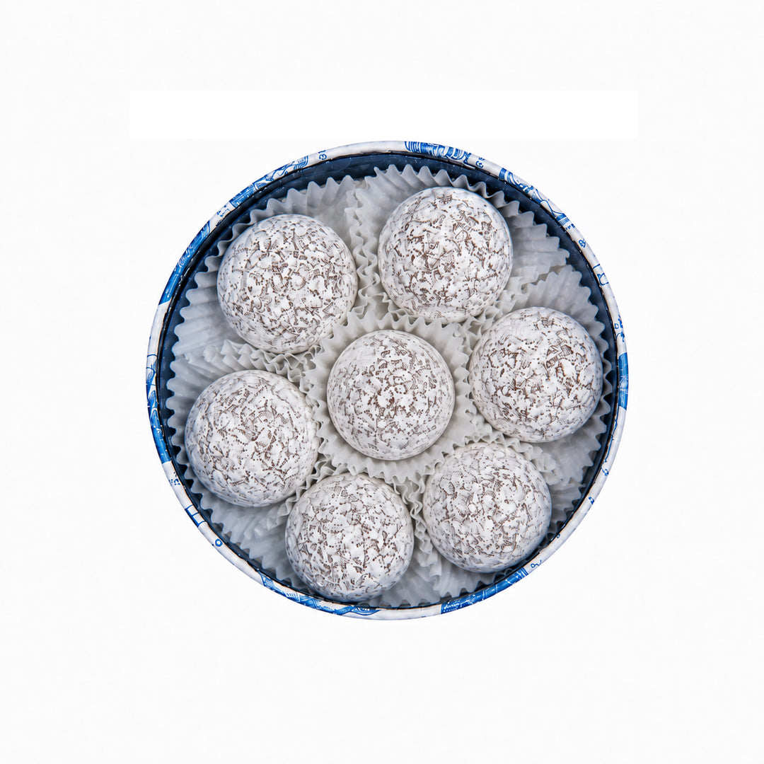 Seven round chocolate-covered confection balls coated in white chocolate or coconut, arranged in a circular white paper liner inside a round blue and white patterned box, on a plain white background.